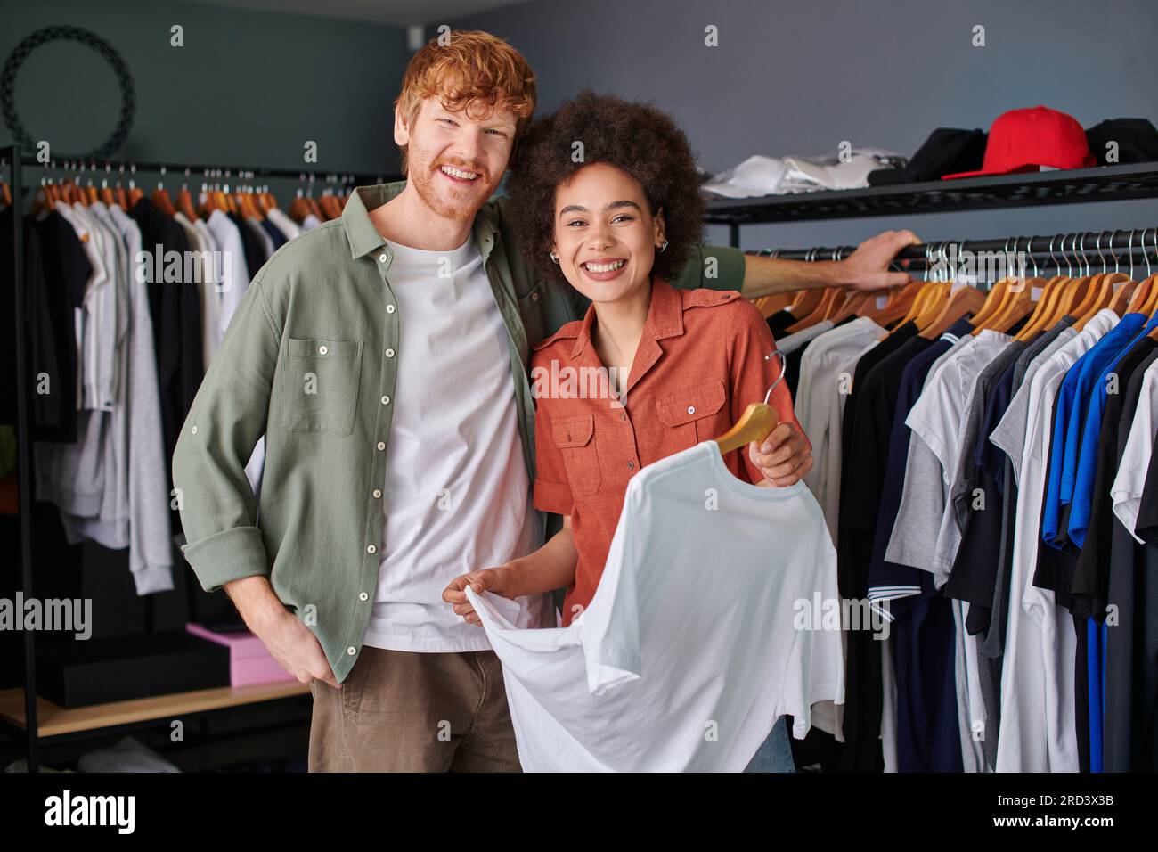 Positif jeune artisan multiethnique regardant la caméra tout en se tenant près de vêtements sur des cintres et travaillant dans un studio d'impression, jeune petite entreprise possède Banque D'Images