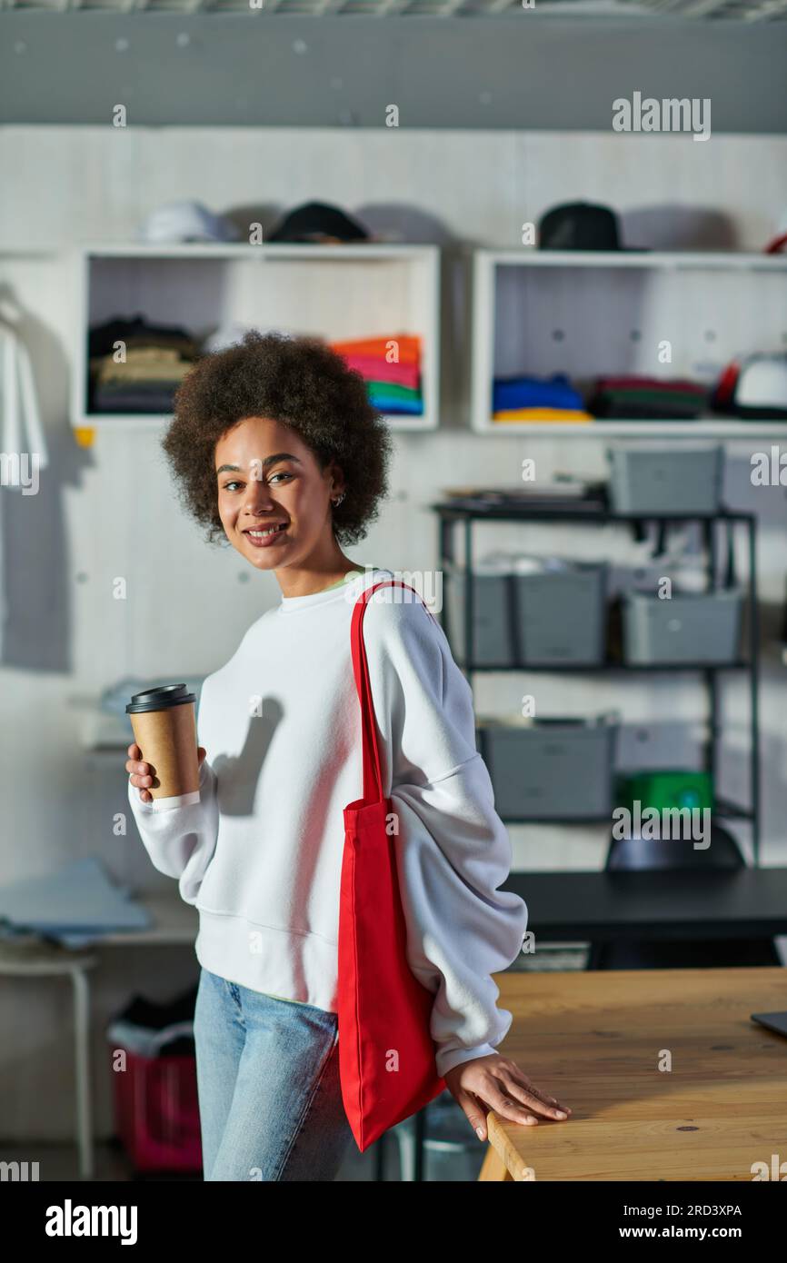 Souriant jeune artisan afro-américain avec sac à bandoulière tenant le café pour aller et regardant la caméra près de la table dans le studio d'impression flou, passionné Banque D'Images