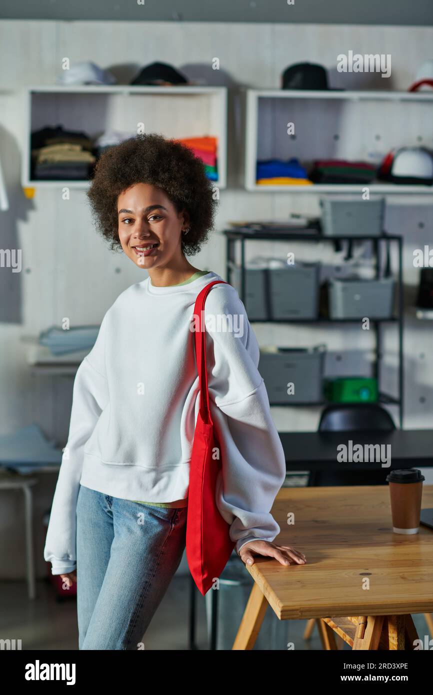 Positif jeune artisan afro-américain avec sac à bandoulière regardant la caméra et debout près du café pour aller sur la table dans le studio d'impression, enthousiaste Banque D'Images