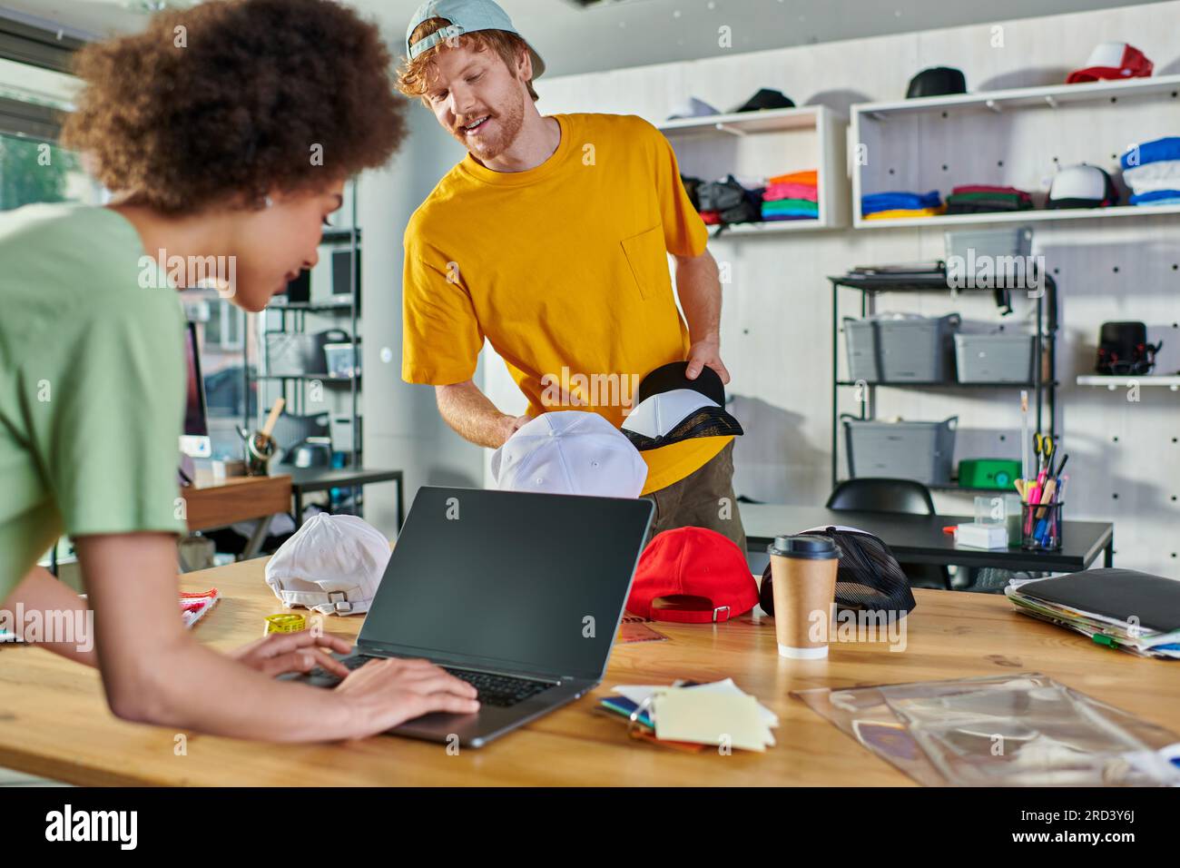 Jeune designer souriant tenant des snapshots tout en parlant à un collègue afro-américain en utilisant un ordinateur portable près des échantillons de couleur dans un studio d'impression, une petite entreprise Banque D'Images