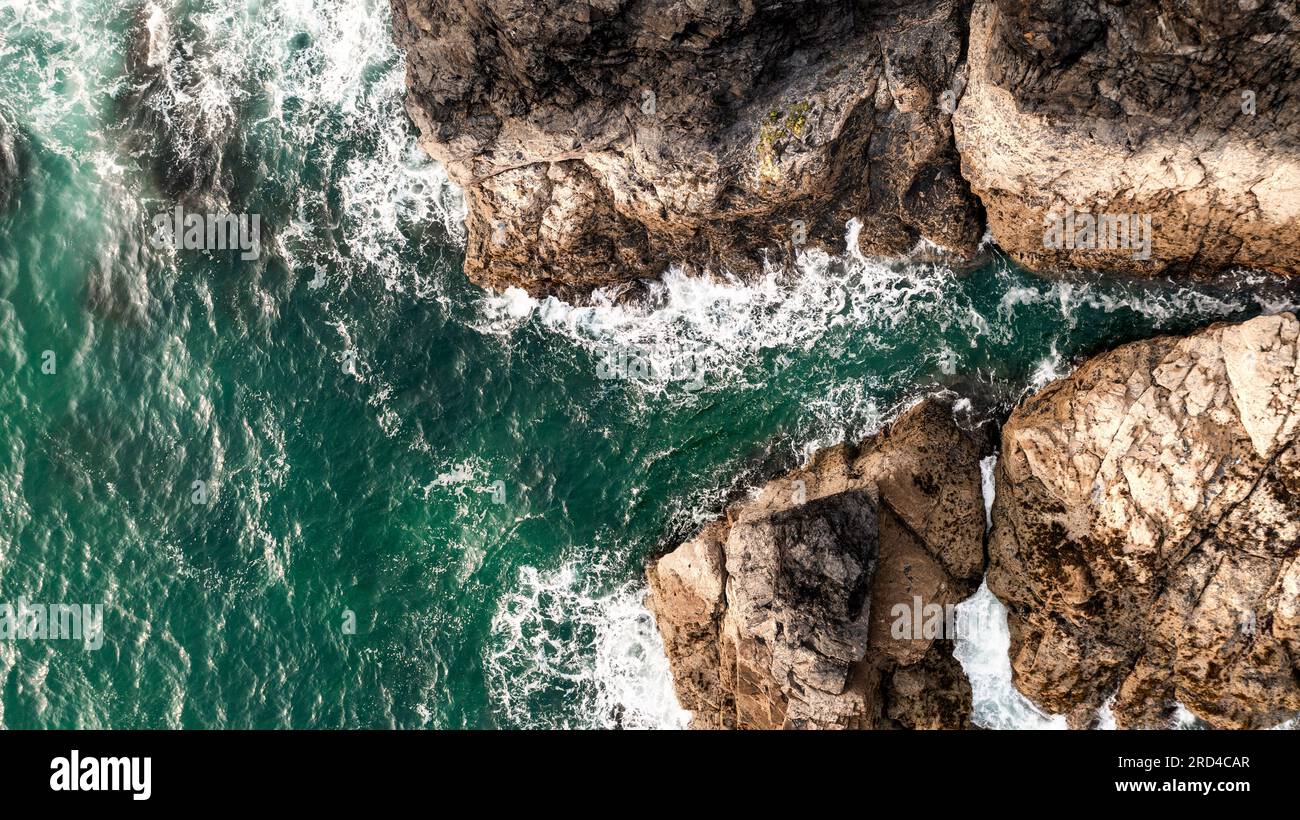 Vue aérienne directement au-dessus des vagues blanches se brisant sur des roches dures de pierre dans un environnement accidenté sur la côte de Cornwall avec espace de copie au-dessus du vert émeraude Banque D'Images