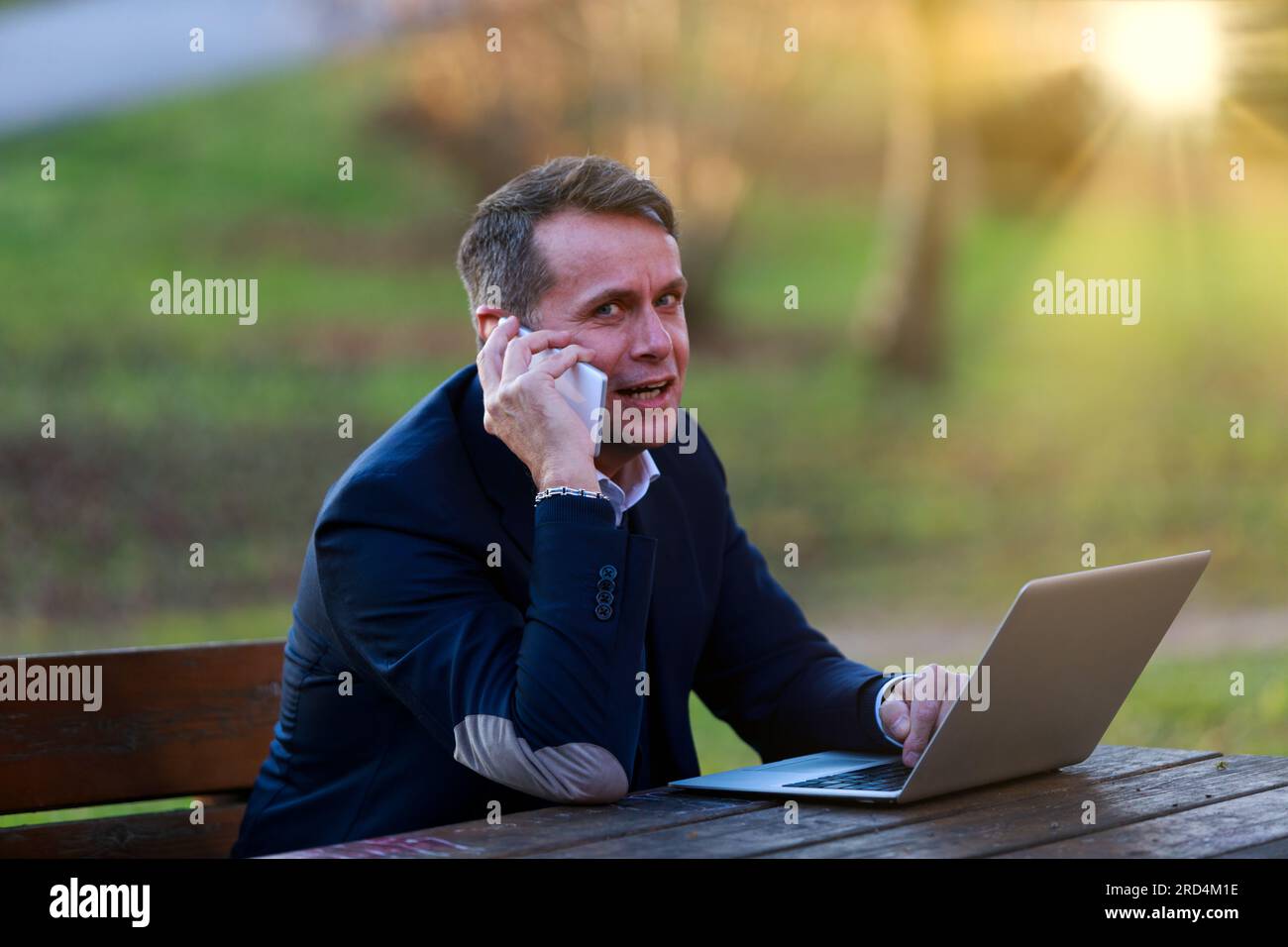 L'homme travaille avec un ordinateur portable et un téléphone dans un parc, toujours accessible mais dans les genoux de la nature. Porte une veste en velours avec des renforts aux coudes, comme une gauche stéréotypée Banque D'Images
