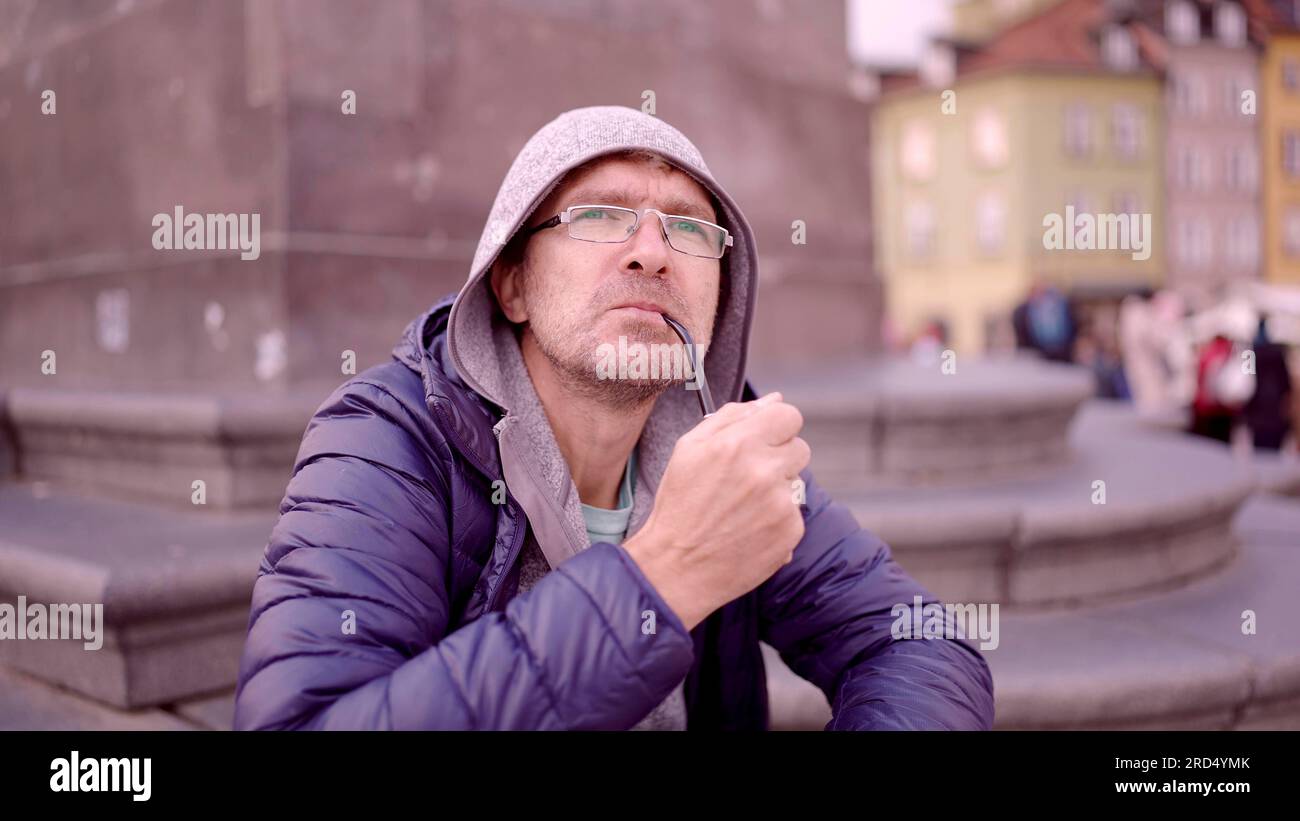 Portrait d'un homme adulte avec des lunettes assis dans la capuche sur la place et fumant un pipe de tabac sur la place du Palais, dans la vieille ville de Varsovie Banque D'Images