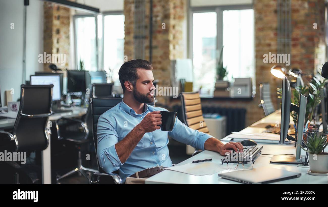 Café frais. Vue latérale d'un jeune homme d'affaires barbu en tenue formelle tenant une tasse de café et regardant l'ordinateur tout en étant assis dans le bureau moderne Banque D'Images
