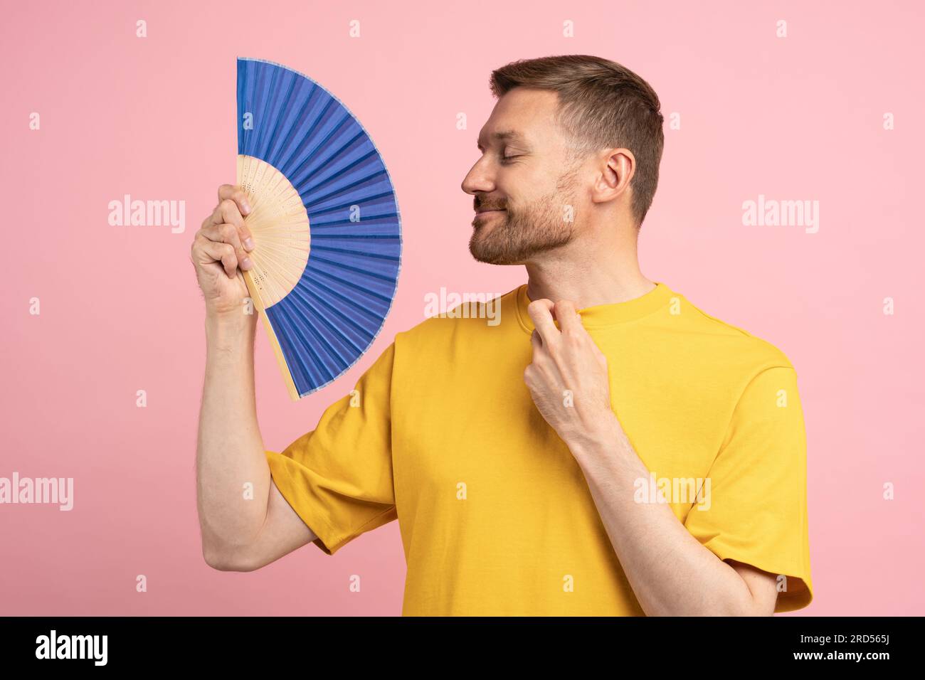 Homme souriant détendu à l'aide d'un ventilateur de papier par temps chaud d'été profitant de l'air frais isolé sur studio rose Banque D'Images