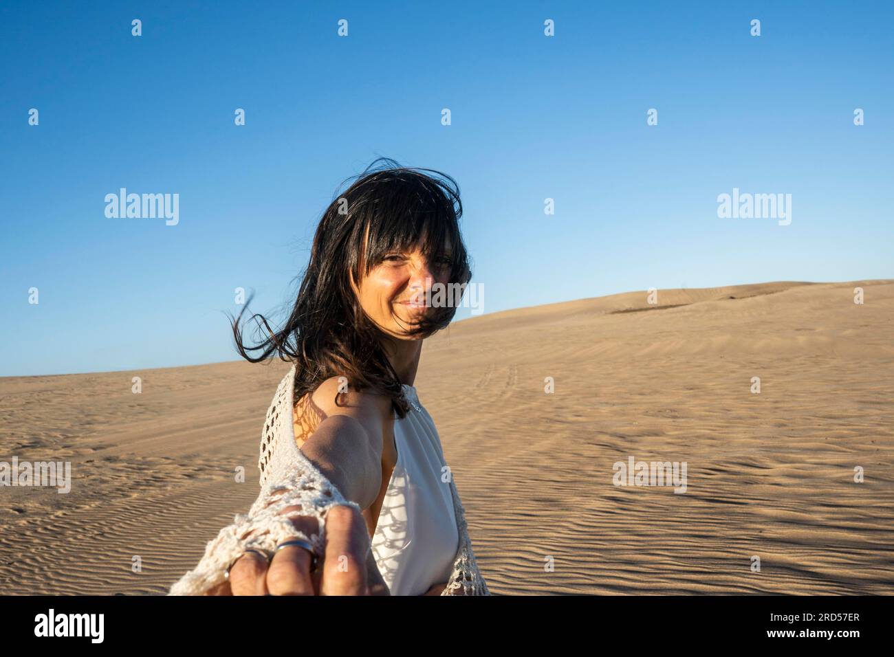 Suivez-moi concept. Une femme souriante aux cheveux longs marchant sur le sable tenant la main de quelqu'un. Banque D'Images