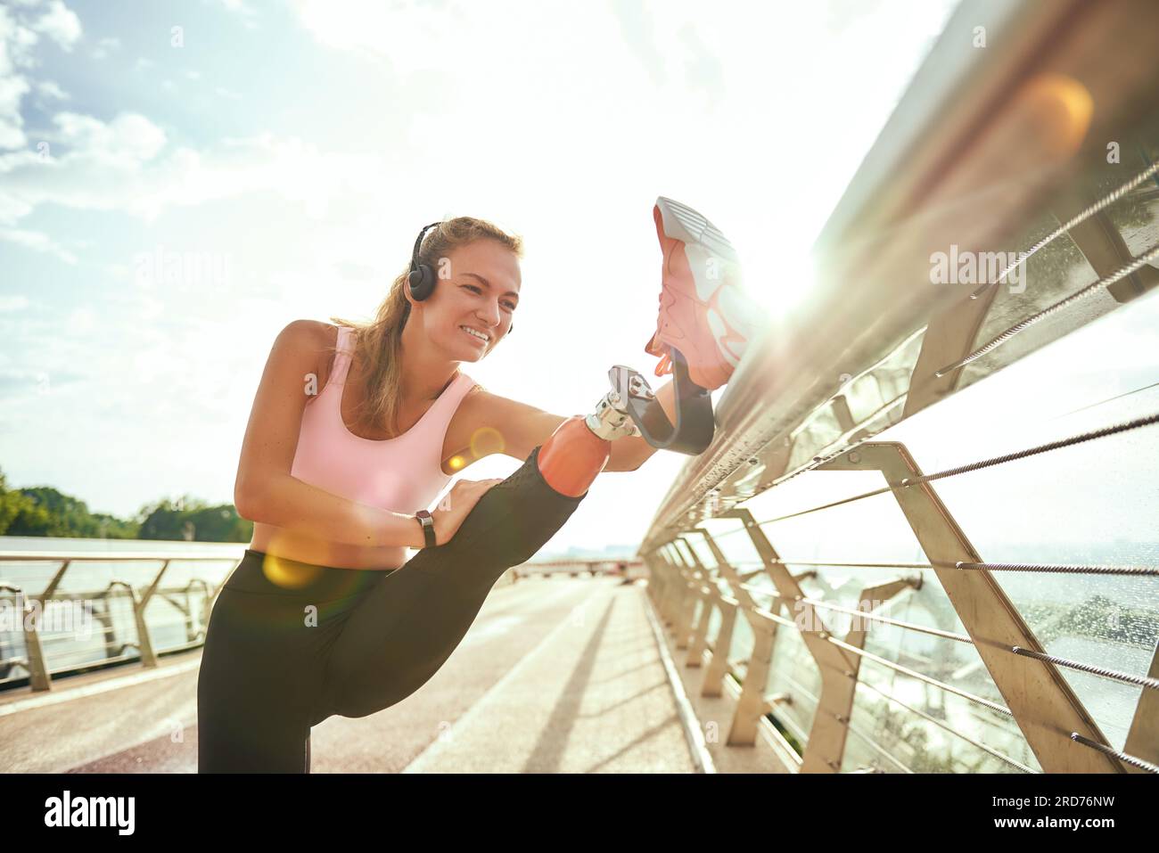 Femme handicapée positive en vêtements de sport et écouteurs étirant la jambe prothétique tout en se tenant debout sur le pont. Écouter de la musique Banque D'Images