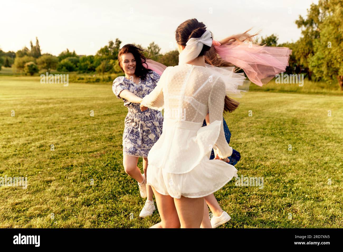 Heureuse mariée charmante dans une robe blanche avec un voile blanc et deux demoiselles d'honneur avec des voiles roses se tenant la main et dansant sur la pelouse dans un parc d'été. CH Banque D'Images