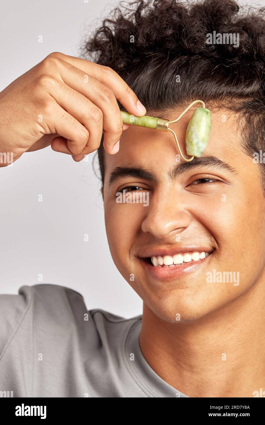 Jeune beau mec souriant utilisant jade dé-souffler le rouleau de visage pour garder sa peau douce et lisse. L'homme swarthy avec les cheveux bouclés noirs maintient son naturel Banque D'Images