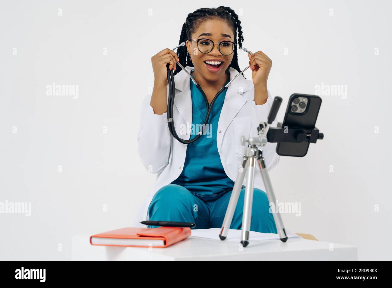 Femme médecin afro-américaine heureuse porte un manteau médical tenant et utilisant un téléphone portable sur fond blanc Banque D'Images