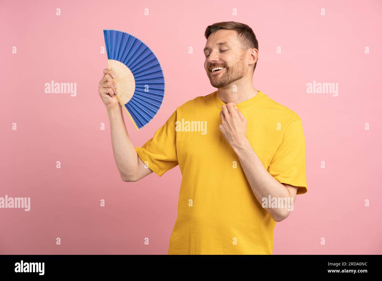 Homme insouciant utilisant le ventilateur de papier au temps chaud d'été appréciant l'air frais isolé sur studio rose Banque D'Images