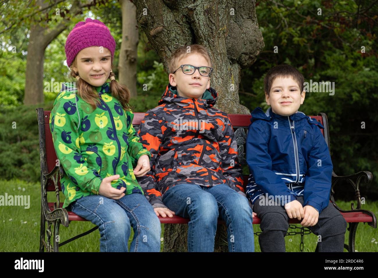 Trois enfants d'âges différents, 7, 10 et 5 ans, sont assis en vestes sur un banc au printemps Banque D'Images
