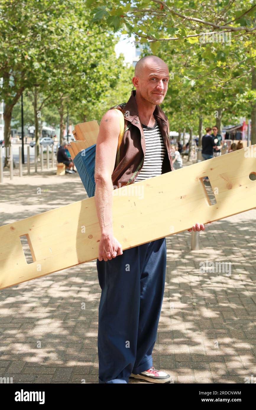 Londres, Royaume-Uni. 20 juillet 2023. 'Bench invasion' de la Compagnie Krak. Dans la performance 'Bench invasion', l'artiste belge Dieter Peter Raphaël Missiaen se déplace dans l'espace public avec dix personnes locales, chacune avec un banc sous le bras, à la recherche de contacts. À un moment inopiné, ils installent des bancs et invitent activement les gens à s’asseoir devant eux. De cette façon, il (re)connecte les gens. Le Festival d'ensemble est un festival de quatre jours de théâtre, de cirque, de danse, de musique et d'art numérique gratuits au Royal Victoria Dock. Ensemble Festival explore les thèmes de l'écologie, de la vie moderne et de la reconnexion, et comprend deux jours de pop-up per Banque D'Images