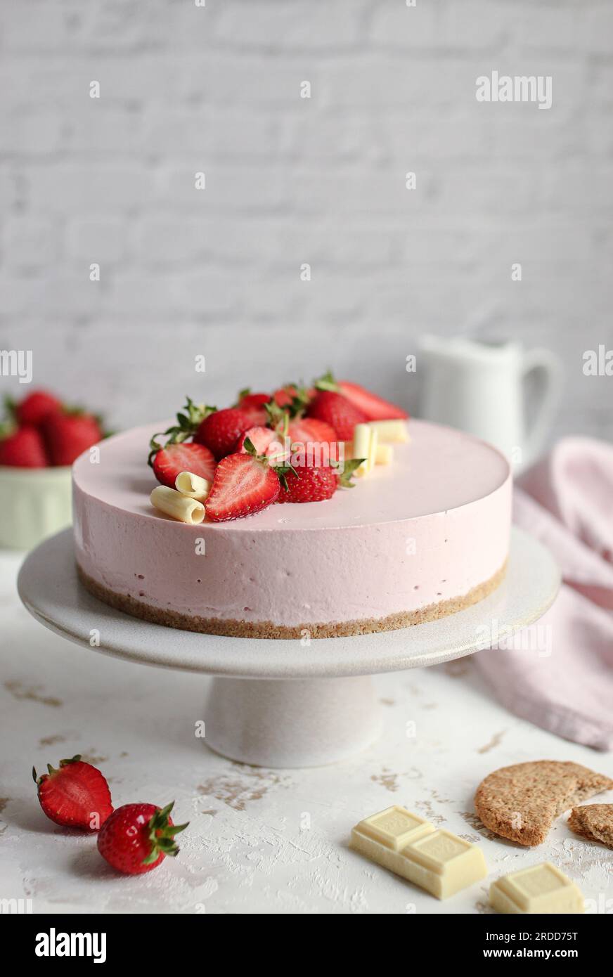 Cheesecake aux fraises sur un stand à gâteaux blanc, décoré avec des fraises fraîches et du chocolat blanc. Serviette rose et pot de lait blanc. Banque D'Images