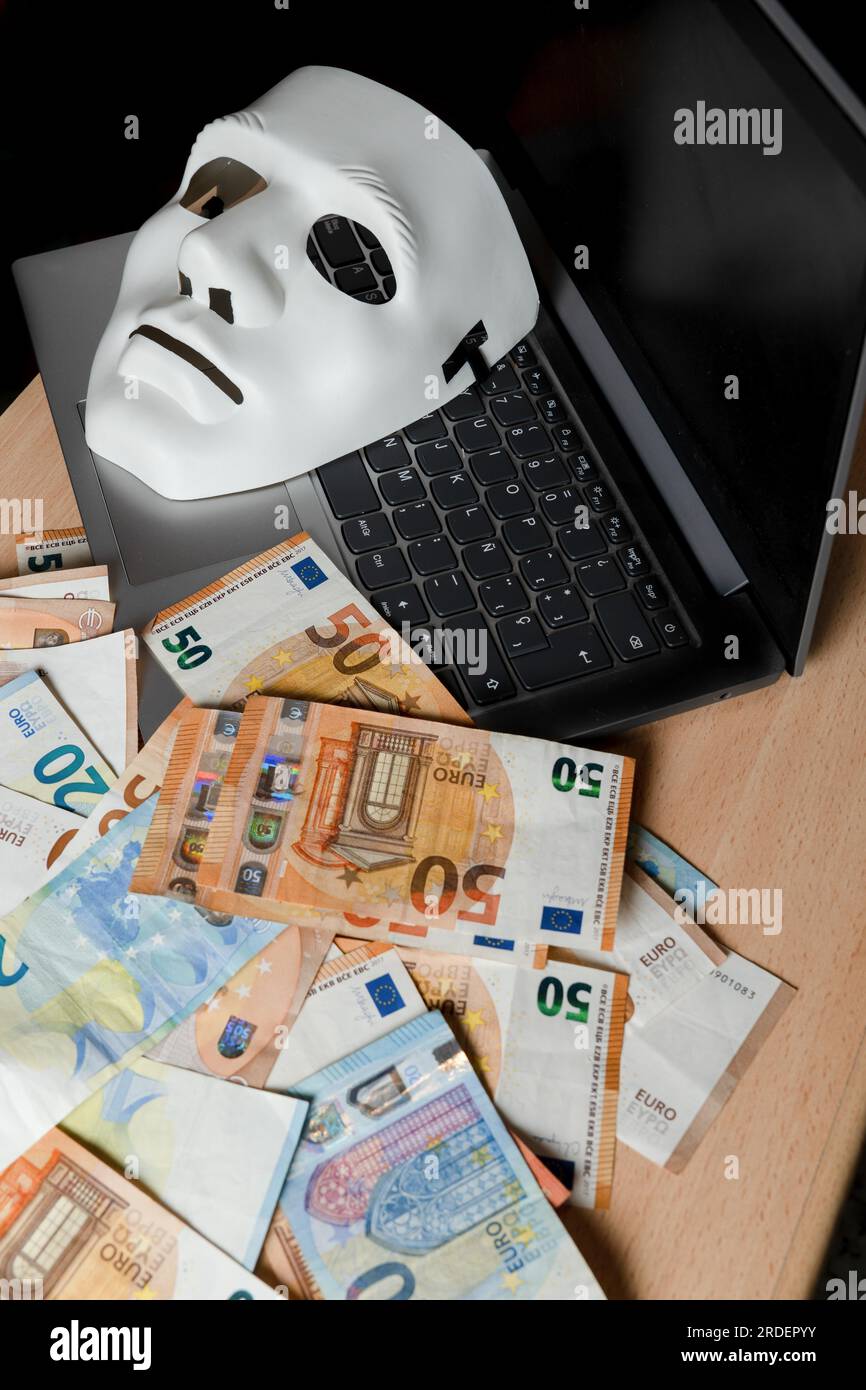 Sur un ordinateur portable, il y a un masque blanc et une pile de billets en euros, concept de cybercriminalité Banque D'Images