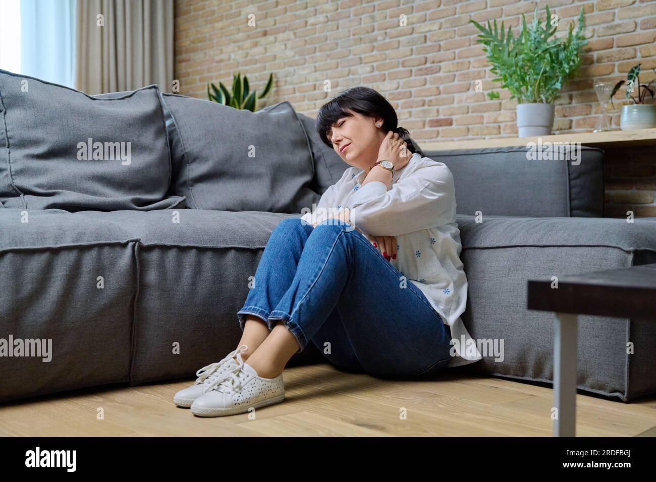 Femme mature souffrant de douleurs au cou, assise sur le sol dans le salon Banque D'Images