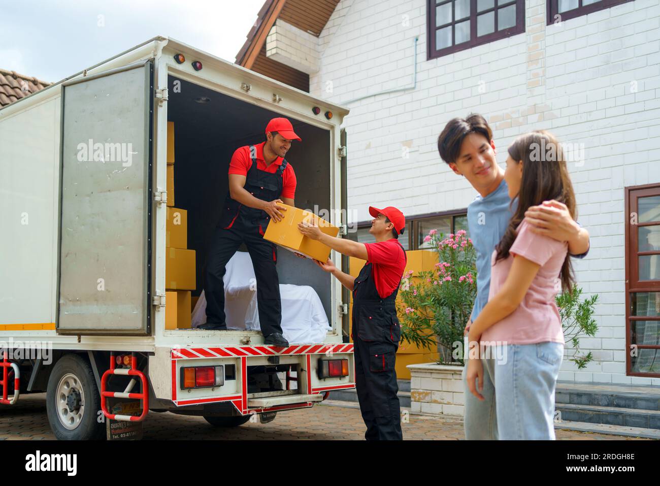 Un couple asiatique se tient ensemble, observant avec anticipation alors que les travailleurs emménagent dans leur nouvelle maison. L'excitation remplit l'air alors qu'ils anticipent avec impatience Banque D'Images