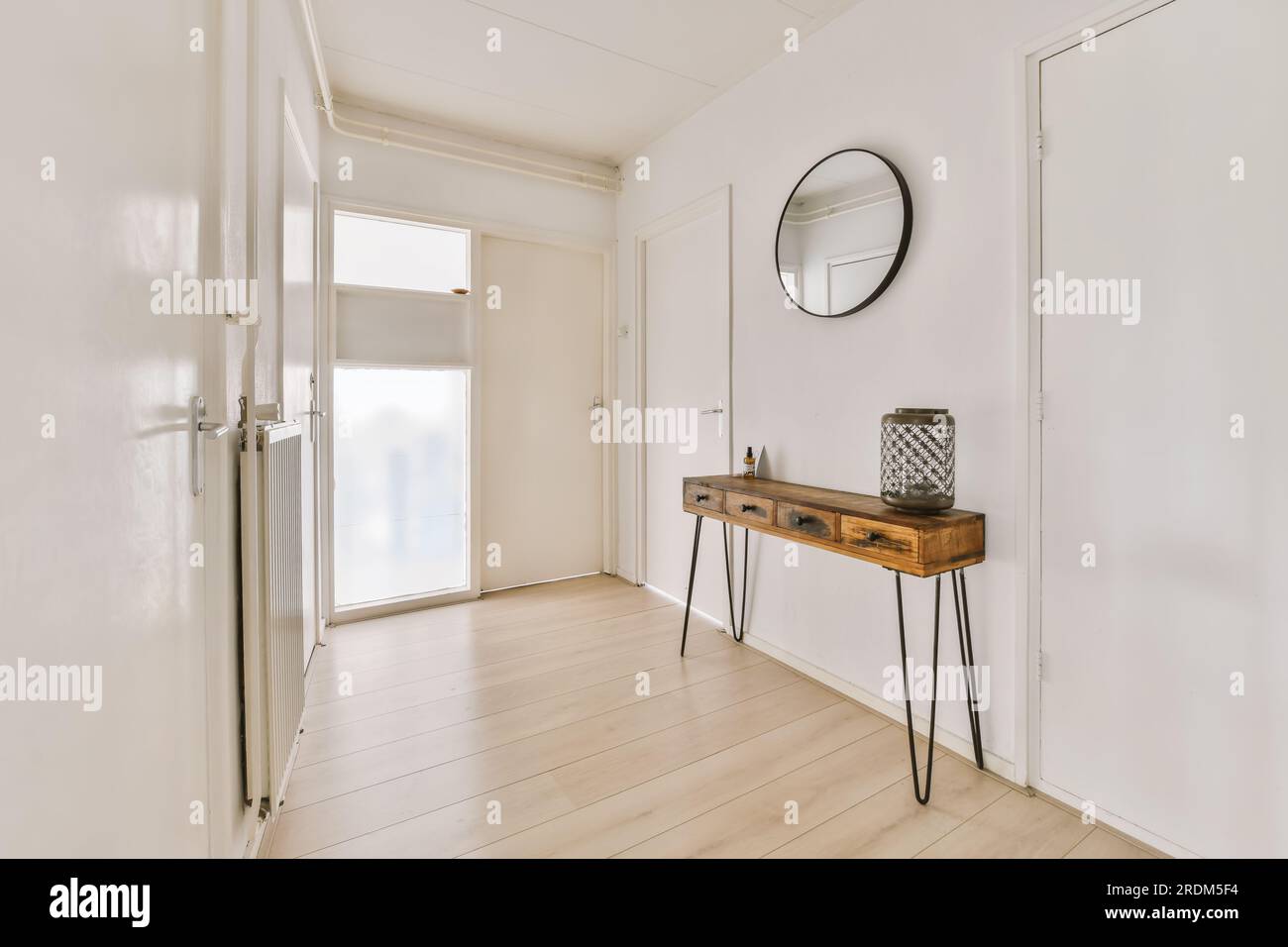 un couloir avec des murs blancs et du parquet la chambre a une table console en bois d'un côté il y a un grand miroir rond Banque D'Images