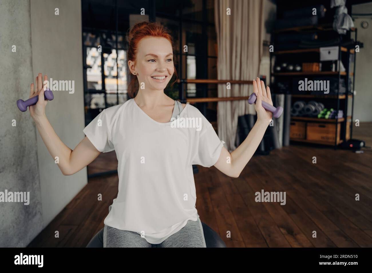 Heureuse femme rousse européenne en tenue active est assise sur le fitball argenté, levant les bras avec des haltères dans le studio de fitness. Vibrations d'entraînement positives ! Banque D'Images