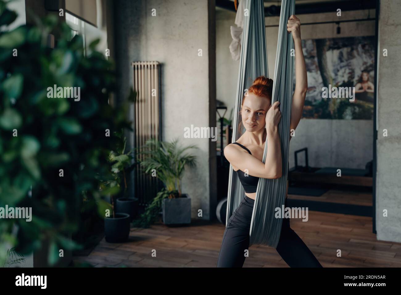Belle femme aux cheveux rouges en tenue active repose après le yoga de mouche dans le studio de fitness. Détente sur un hamac gris, sourire à la caméra. Embrasser la santé, Banque D'Images