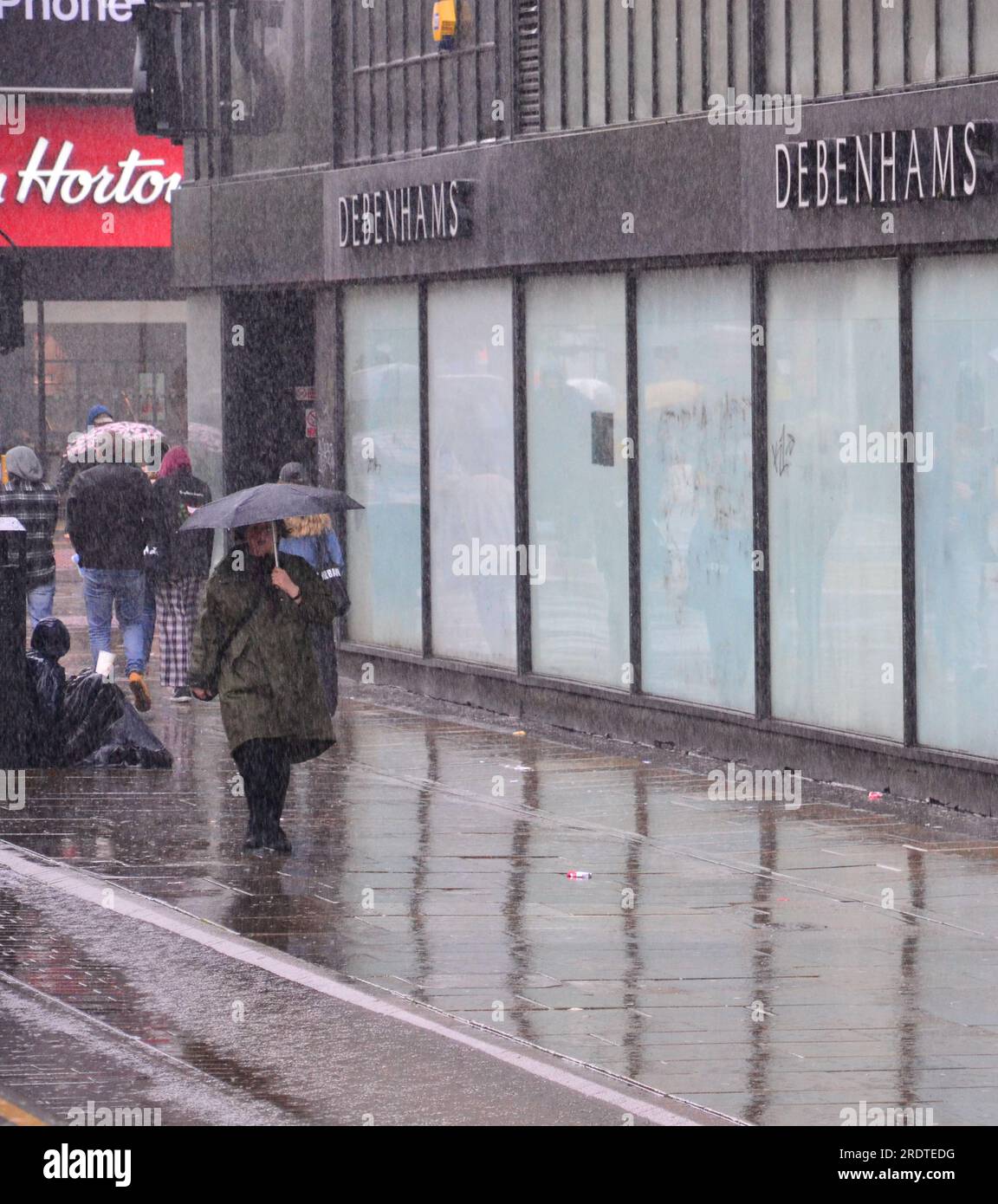 Manchester, Royaume-Uni, 23 juillet 2023. Les acheteurs se battent contre les pluies torrentielles sur Market Street, dans le centre de Manchester, au Royaume-Uni, tandis que le met Office émet un avertissement météorologique jaune pour le nord du pays de Galles, Merseyside, Greater Manchester et Newcastle-upon-Tyne. Des inondations sont probables à certains endroits. Un homme assis appuyé contre la poubelle tient une tasse pour demander de l'argent. Crédit : Terry Waller/Alamy Live News Banque D'Images