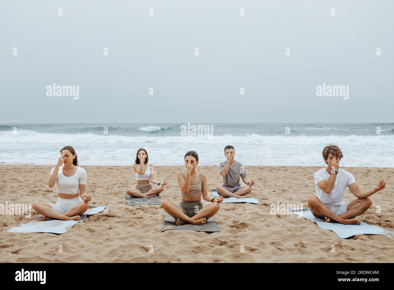 Groupe de femmes et d'hommes pratiquant le yoga sur la plage, touchant leur front avec les doigts, assis sur des nattes sur le sable Banque D'Images