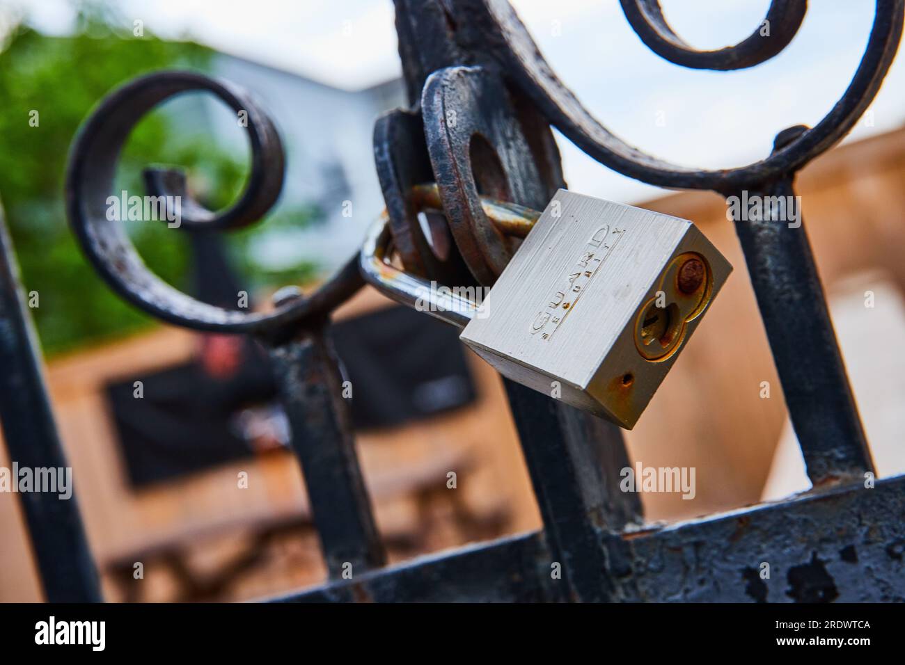 Cadenas de sécurité de garde sur clôture en métal noir rouillé avec bâtiment flou et arbre en arrière-plan Banque D'Images