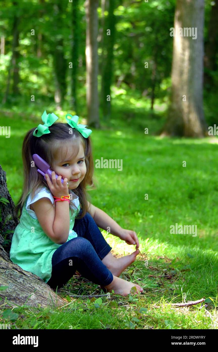 Adorable petite fille prétend avoir une conversation téléphonique. Elle écoute et regarde la caméra. Elle a des queues de porc et des noeuds de cheveux verts. Banque D'Images