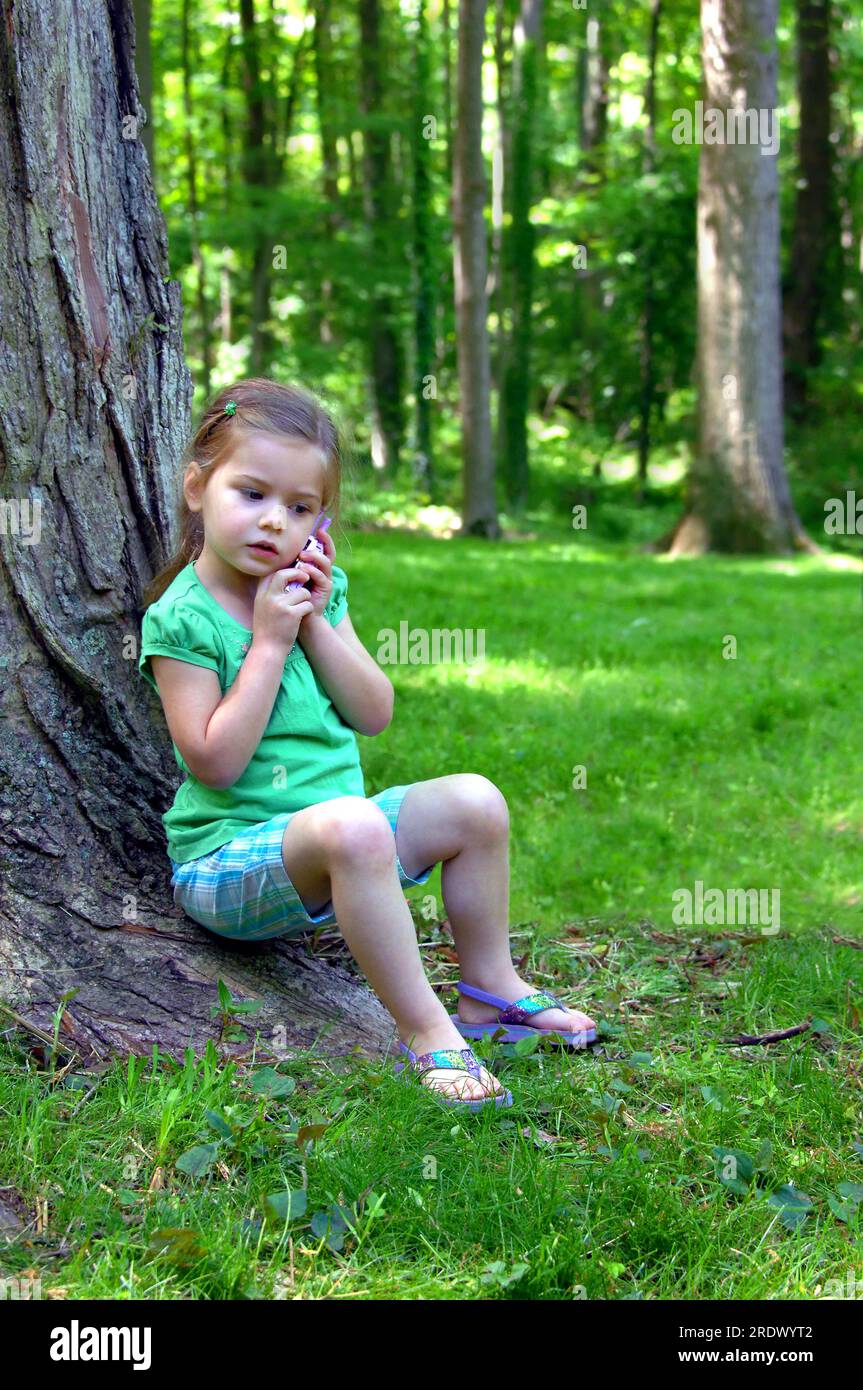Jolie petite fille répond à son téléphone de jeu. Elle est assise à l'extérieur près d'un arbre. Elle porte des tongs. Banque D'Images