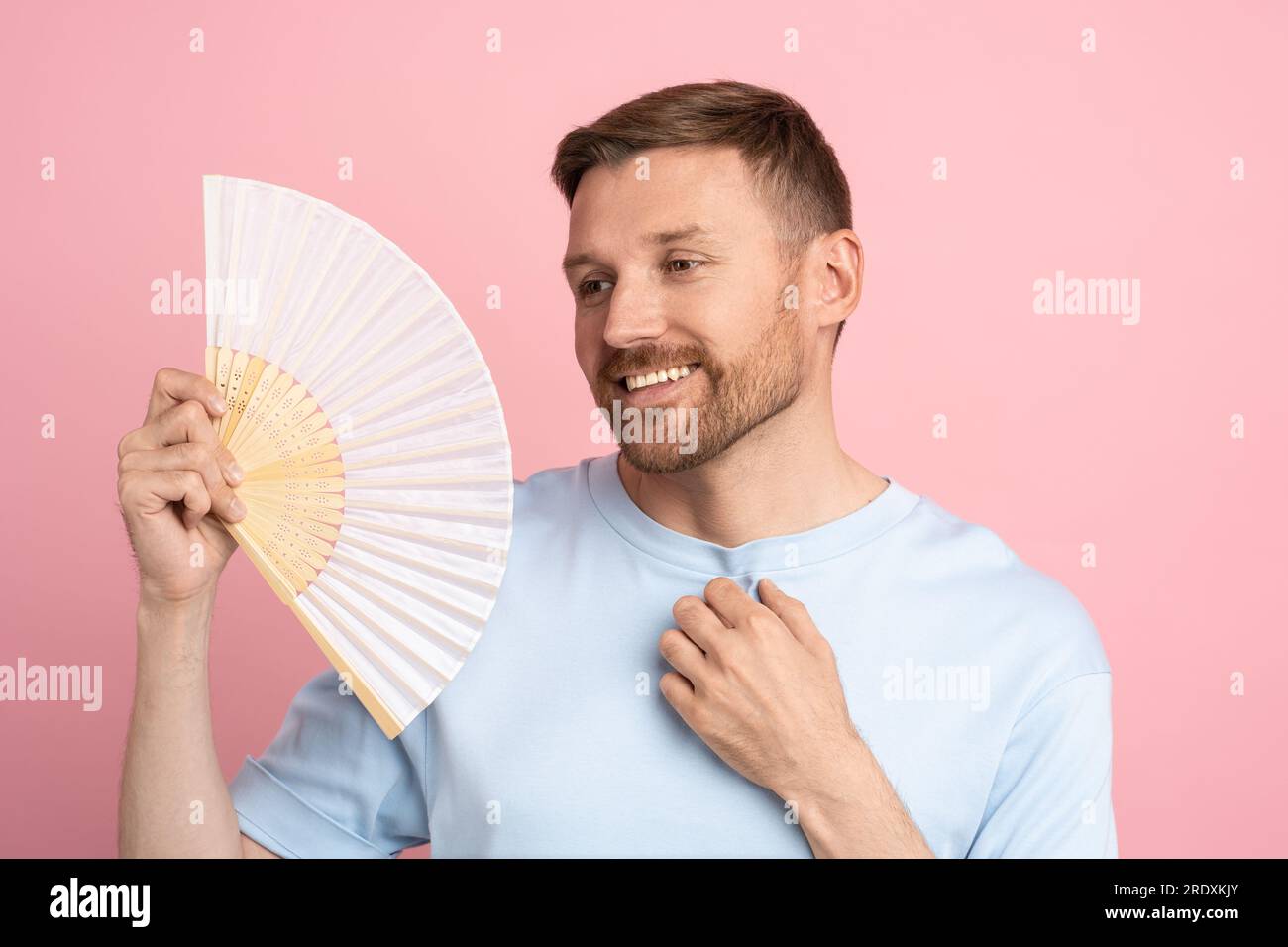 Homme insouciant utilisant le ventilateur de papier au temps chaud d'été appréciant l'air frais isolé sur studio rose Banque D'Images