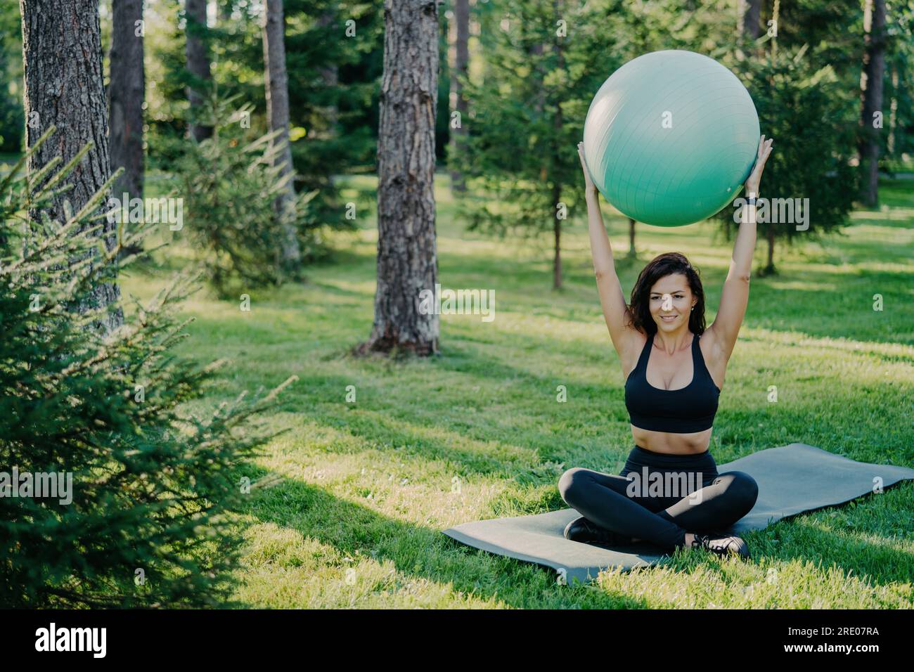 Mince brunette soulève le ballon de fitness au-dessus de la tête, assis en lotus pose sur karemat dans la forêt. Porter des vêtements actifs, pratiquer le yoga à l'extérieur, profiter d'un frais Banque D'Images