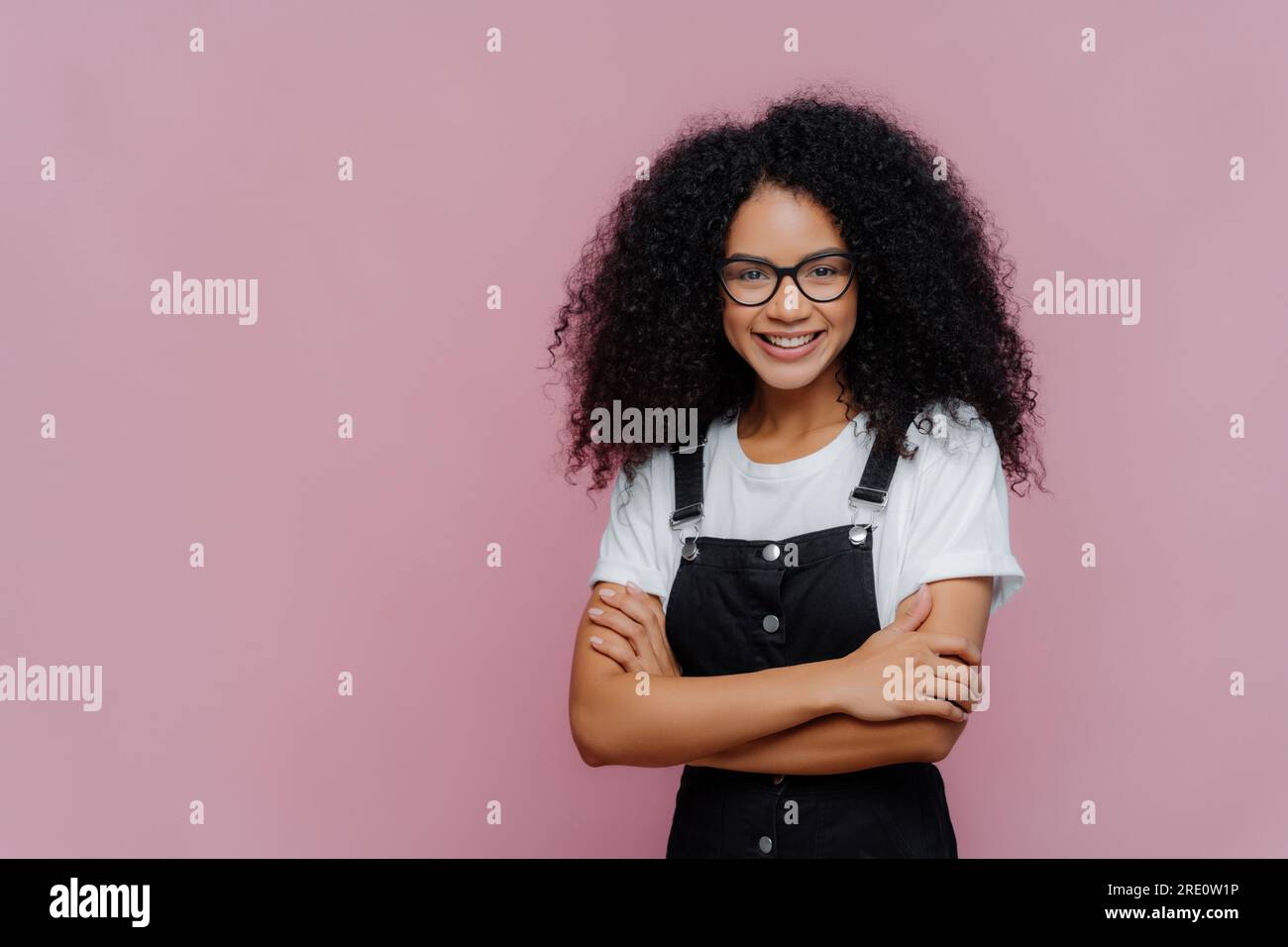 Adolescente heureuse avec afro, lunettes, t-shirt blanc, salopette noire, debout sur fond violet. Banque D'Images