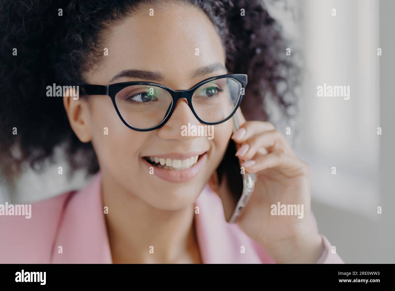 Femme à la peau sombre avec des lunettes sourit, tient le téléphone, discute des investissements avec le partenaire, regarde de côté. Banque D'Images