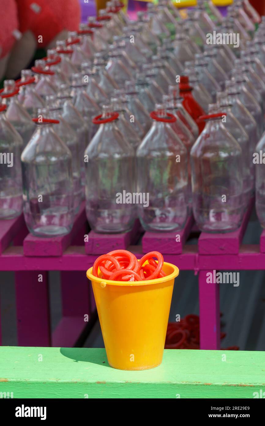 Jeu de lancer d'anneau de carnaval avec une tasse jaune pleine d'anneaux rouges au premier plan, et des rangées de bouteilles en arrière-plan Banque D'Images