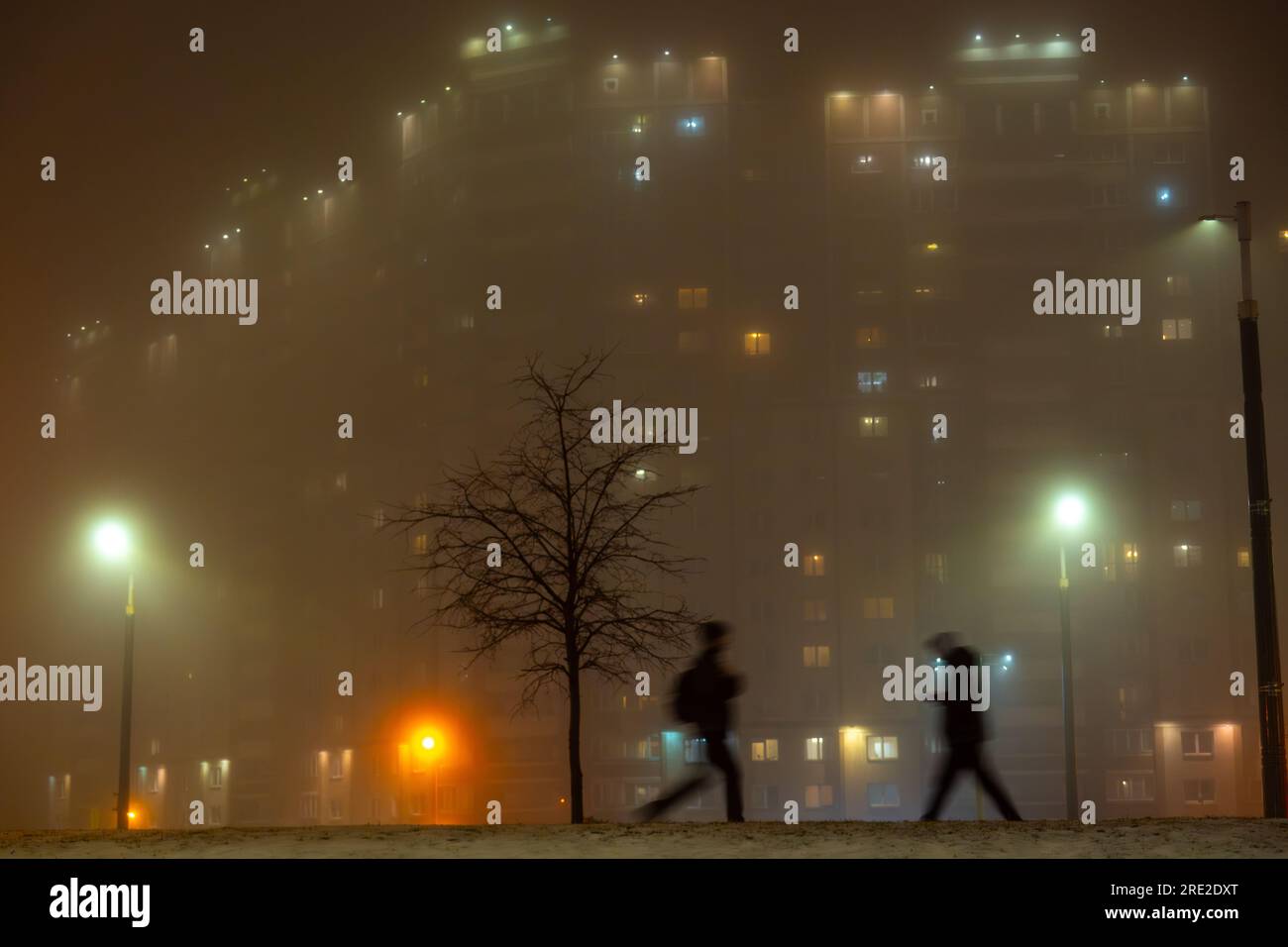 Silhouettes floues de piétons marchant dans la rue avec des lampadaires et un immeuble résidentiel dans un brouillard dense la nuit Banque D'Images