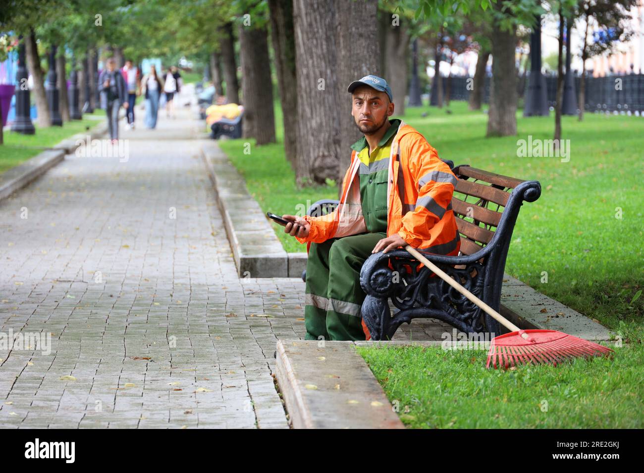 Homme concierge assis avec smartphone sur un banc dans le parc d'été. Travailleur des services publics pendant le repos, nettoyage des rues Banque D'Images