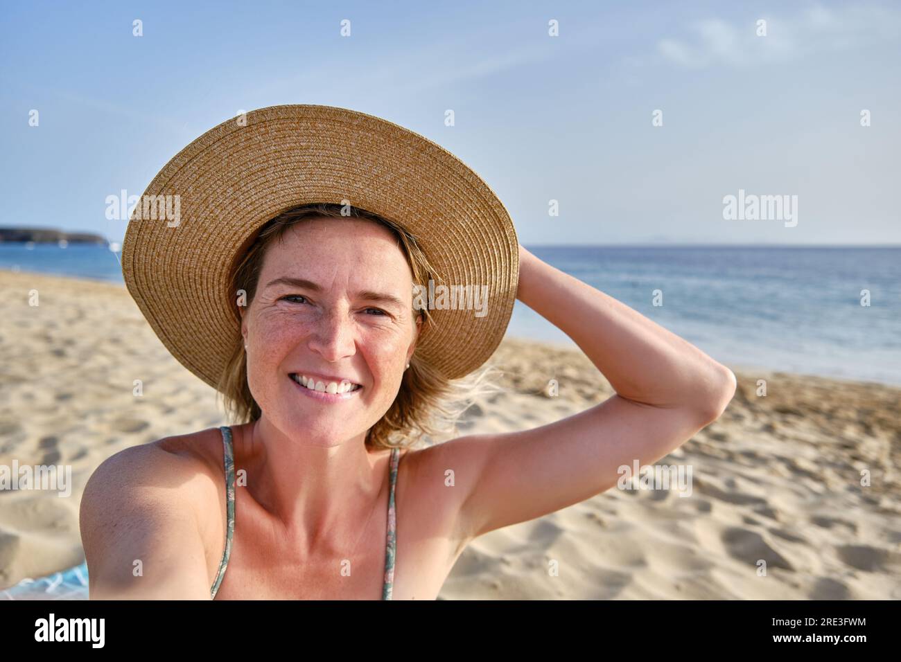 Femme souriante dans le chapeau debout sur la plage de sable et prenant selfie Banque D'Images