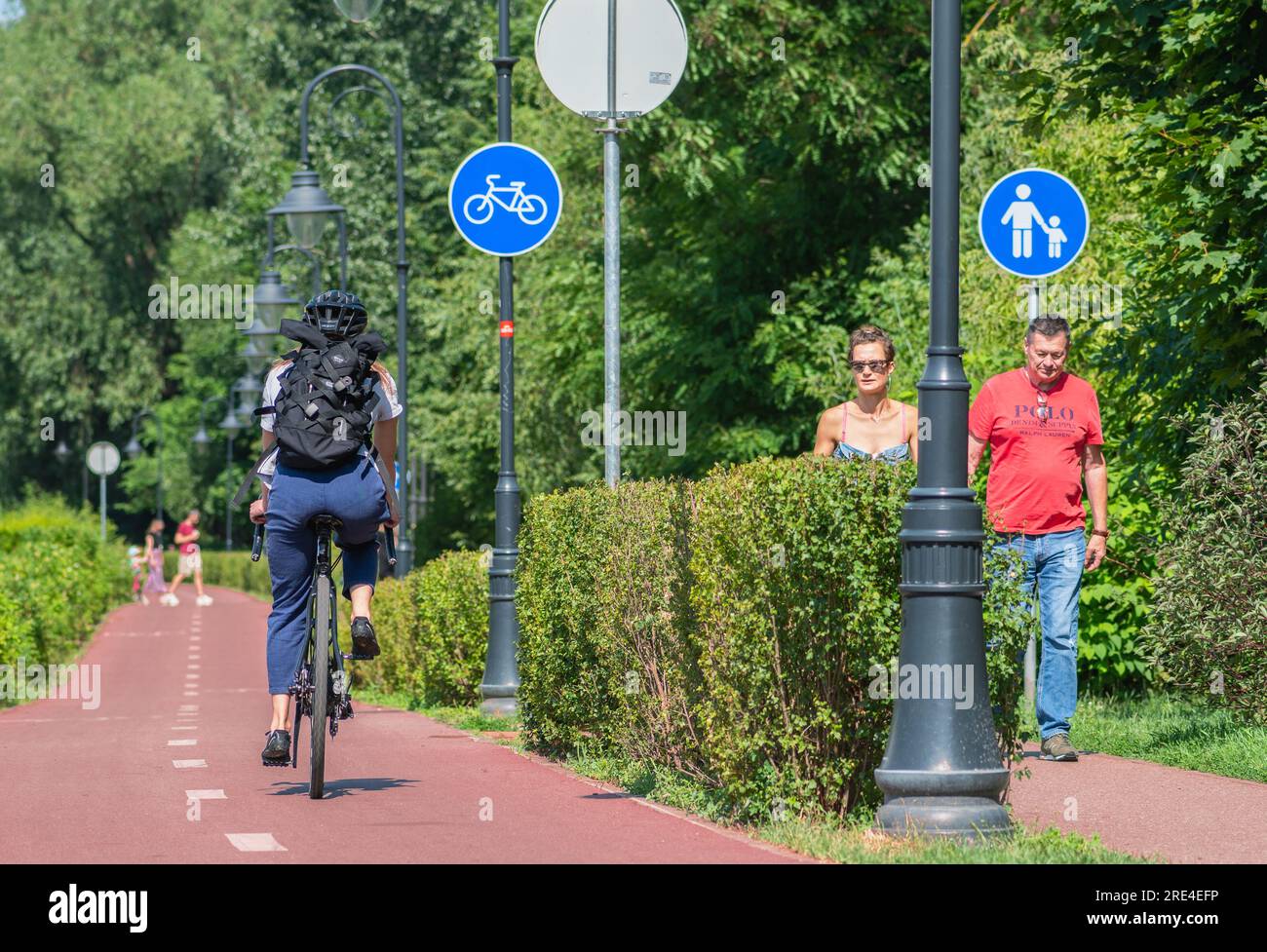 Fille avec casque et sac à dos à vélo sur la route cyclable et couple de piétons marchant sur la passerelle avec panneau bleu ou signal de piste cyclable Banque D'Images