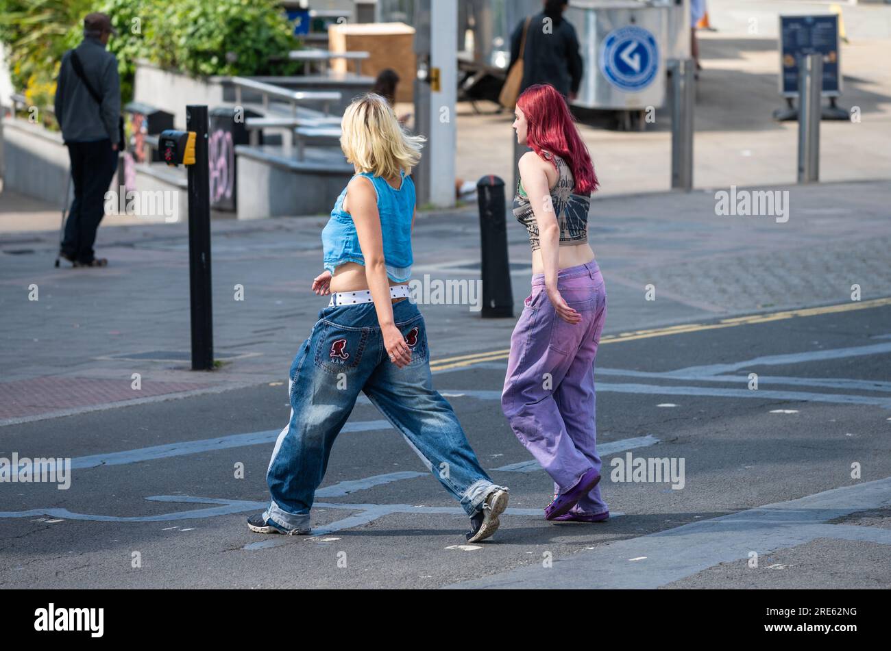 Paire de jeunes femmes minces, filles ou adolescentes, habillées en jeans, marchant à travers une route et faisant du shopping en été, Royaume-Uni. Amis dans les magasins. Banque D'Images