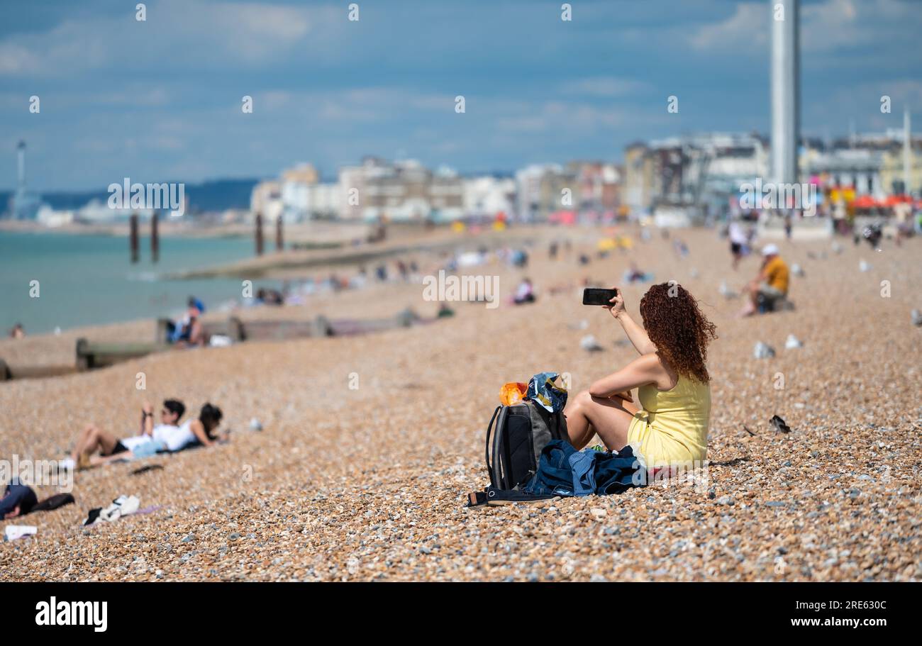 Femme prenant selfie avec smartphone, assise sur la plage de Brighton par une journée nuageuse en été à Brighton & Hove, East Sussex, Angleterre, Royaume-Uni. Banque D'Images