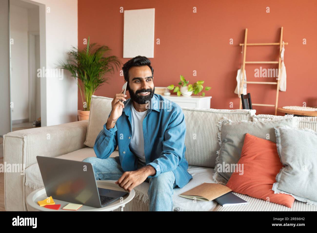 Homme indien ayant une conversation téléphonique agréable assis près de l'ordinateur portable à l'intérieur Banque D'Images