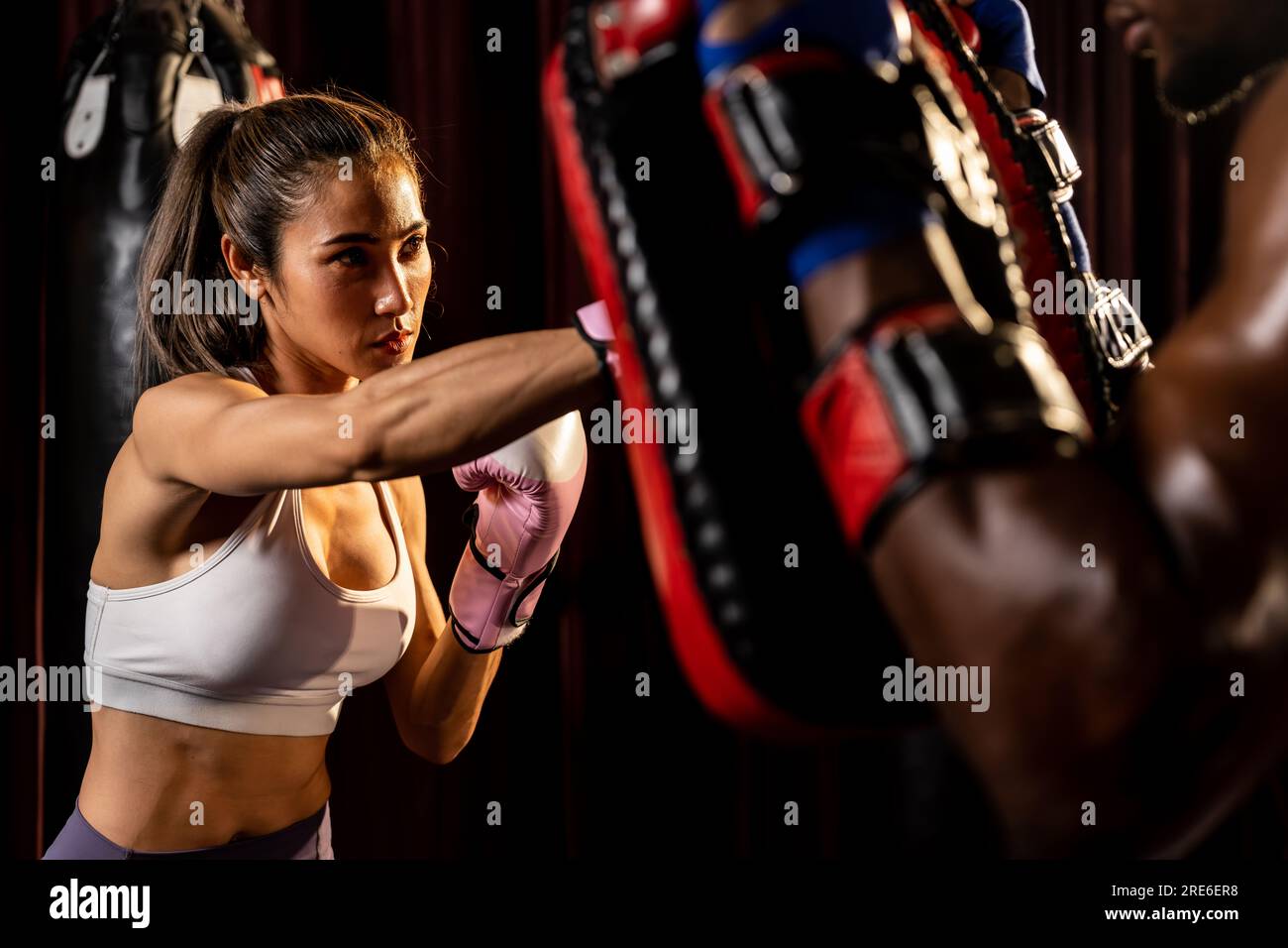 Une femme asiatique, Muay Thai boxer, qui pointait dans une session d'entraînement de boxe féroce, donne la grève à son entraîneur de combat portant des mitaines de poinçonnage, en présentant Banque D'Images