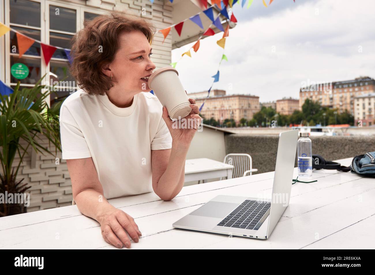 Femme mature assise dans le café en plein air à la table avec ordinateur portable et tasse de papier de café. Brunette d'âge moyen en t-shirt blanc fonctionne et joue en utilisant mod Banque D'Images