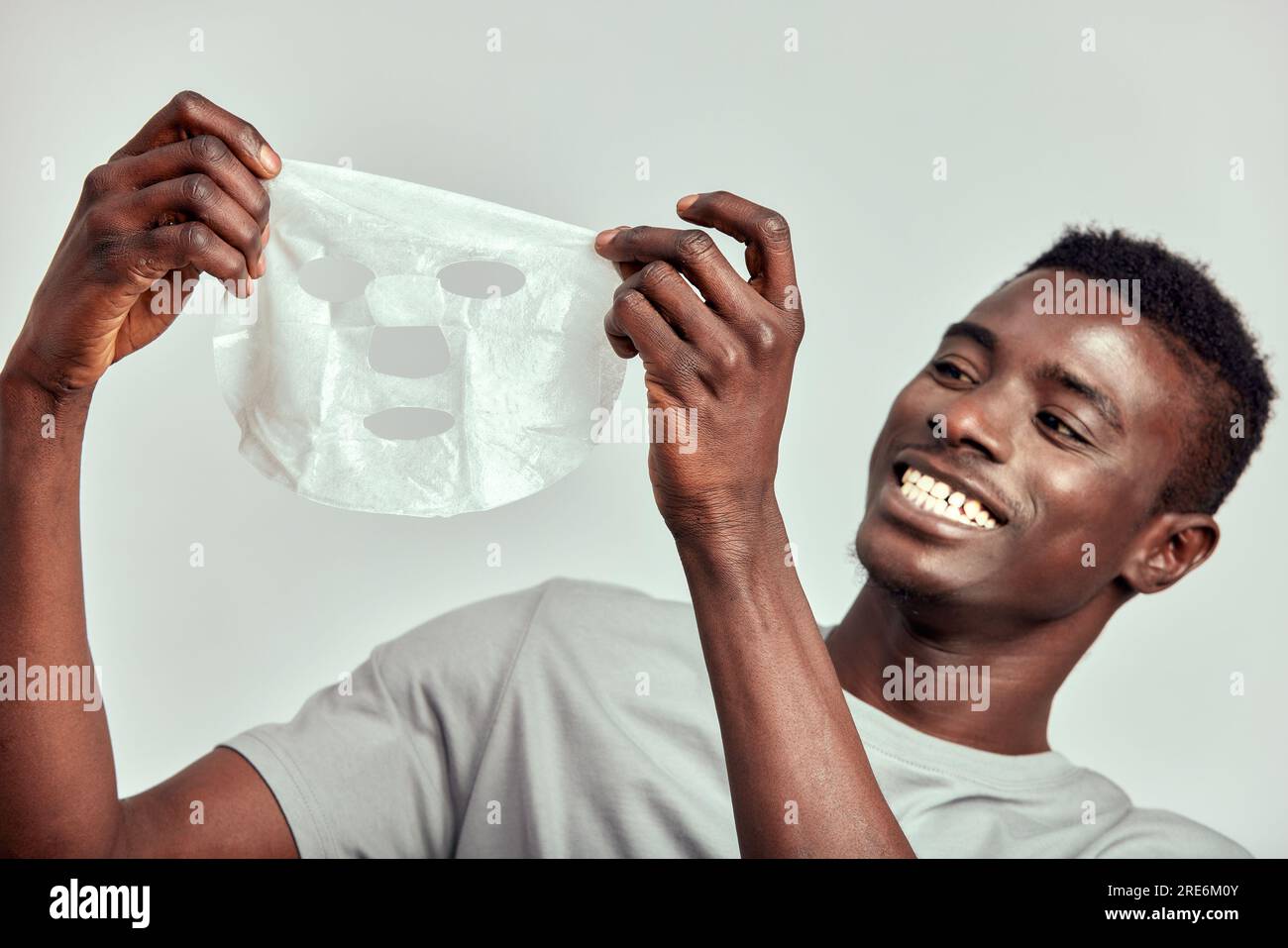 Portrait en studio d'un jeune homme noir souriant appliquant un masque en papier hydratant sur son visage. Heureux homme afro-américain Millennial utilise des produits de beauté dans Banque D'Images
