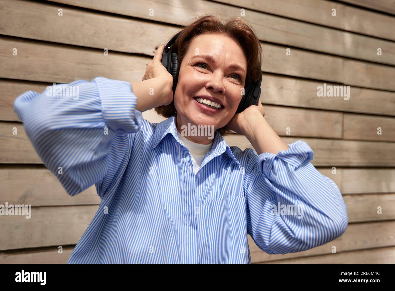 Portrait de femme mature attrayante avec des écouteurs sans fil sur le fond de mur de maison de parement. Femme brune d'âge moyen dans une chemise bleue listenin Banque D'Images
