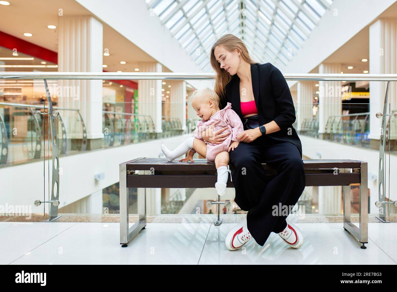 Jeune jolie mère avec un petit bébé est assise sur un banc dans un centre commercial. Maman et fille dans des vêtements roses se détendre et s'amuser tout en faisant du shopping i Banque D'Images