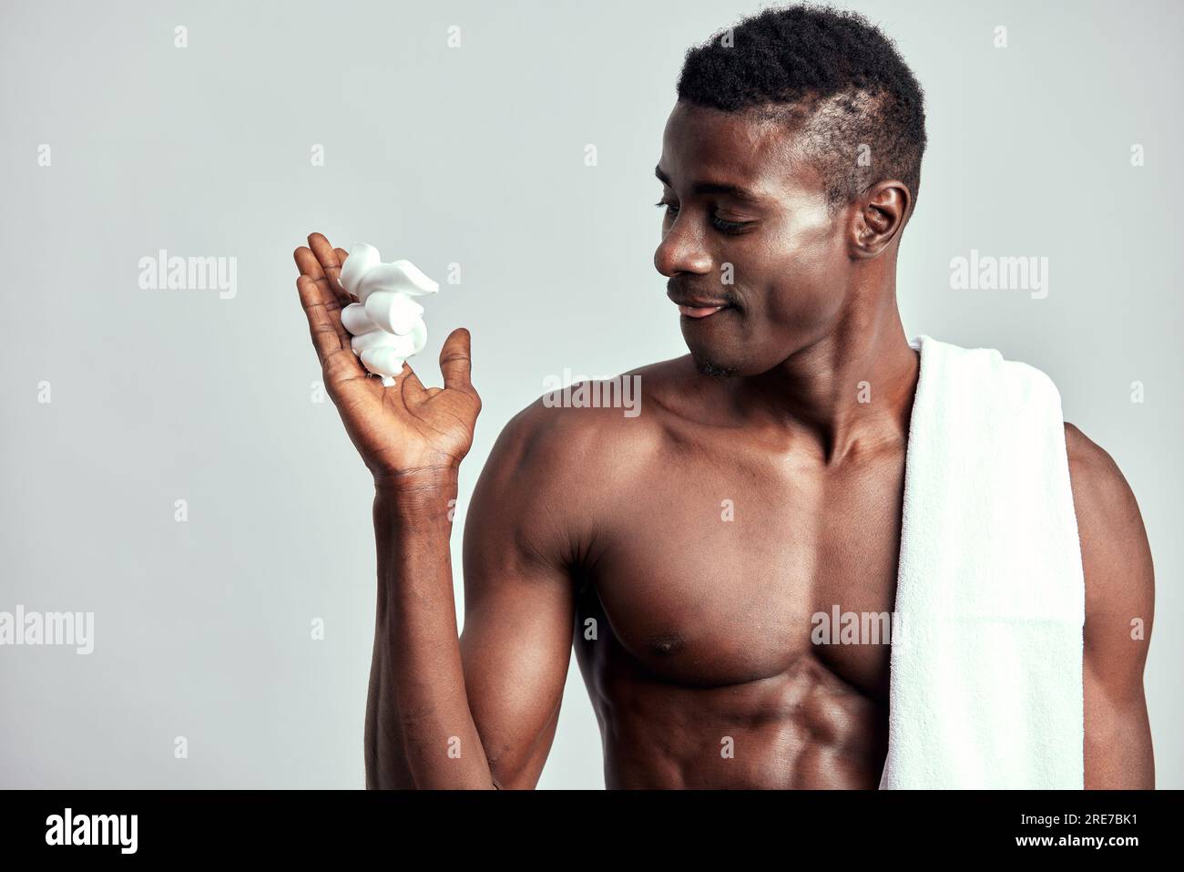 Portrait de studio de beau gars noir avec le torse nu appliquant de la crème sur son visage. Homme afro-américain avec serviette sur son épaule utilise des produits de beauté i Banque D'Images