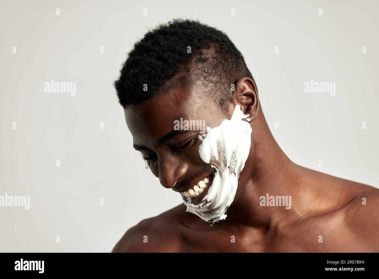 Gros plan portrait studio de beau mec noir souriant avec le torse nu et la crème sur son visage. L'homme afro-américain utilise des produits de beauté dans ses soins de la peau Banque D'Images