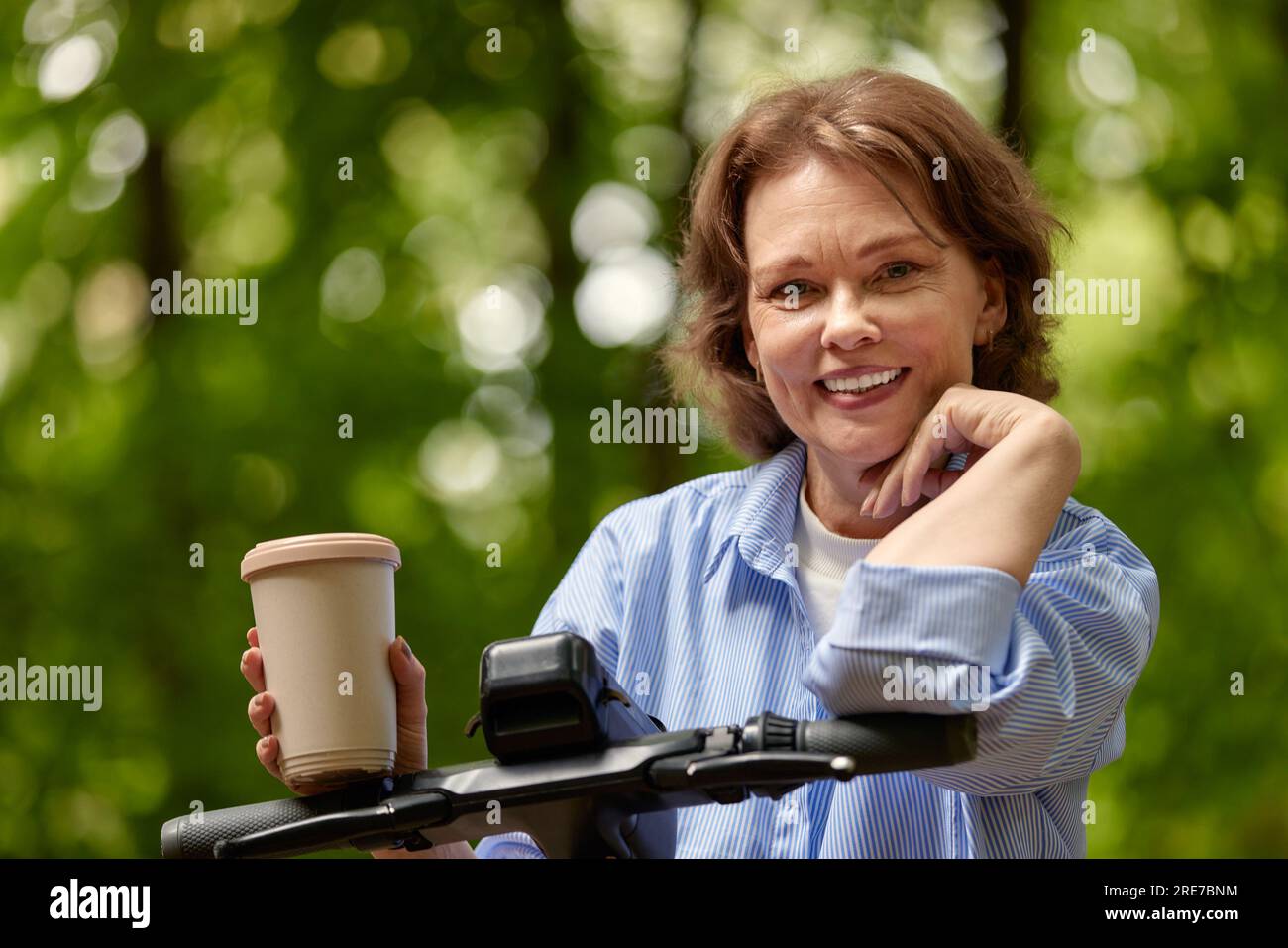 Adorable femme mature avec scooter électrique et tasse à café en papier dans le parc. Joyeuse souriante dame d'âge moyen mène une vie active. Age inclusivi Banque D'Images