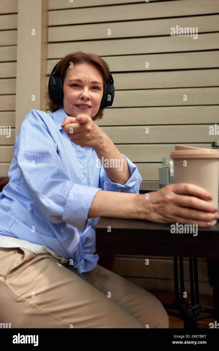 Adorable femme mature avec des écouteurs sans fil assis à une table dans un café en plein air. Brunette souriante d'âge moyen en chemise bleue avec tasse à café en papier po Banque D'Images